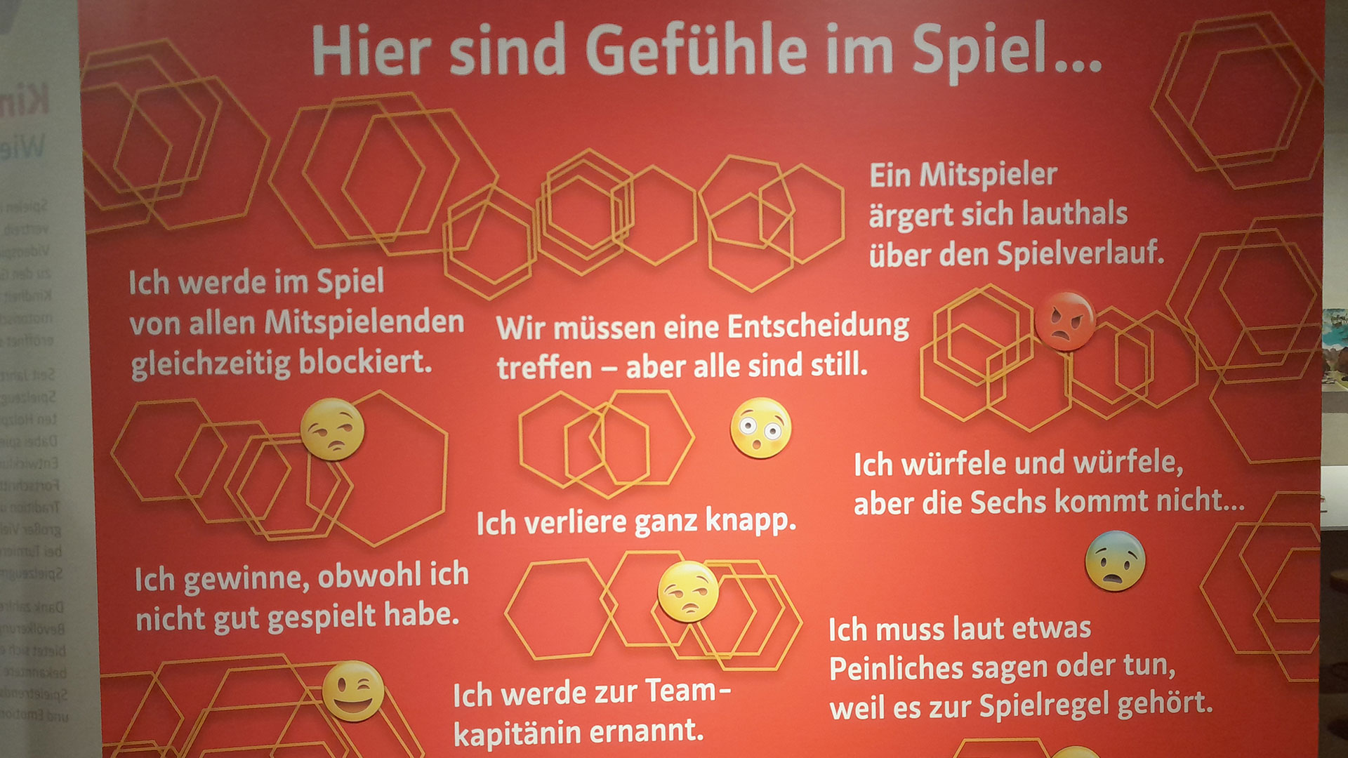 Eine deutsche Tafel mit dem Titel Hier sind Gefühle im Spiel... vor rotem Hintergrund und mit gelben Sechsecken, auf der verschiedene spielbezogene Emotionen aufgeführt sind, jeweils mit einem entsprechenden Emoji-Gesicht in weißer und gelber Schrift.