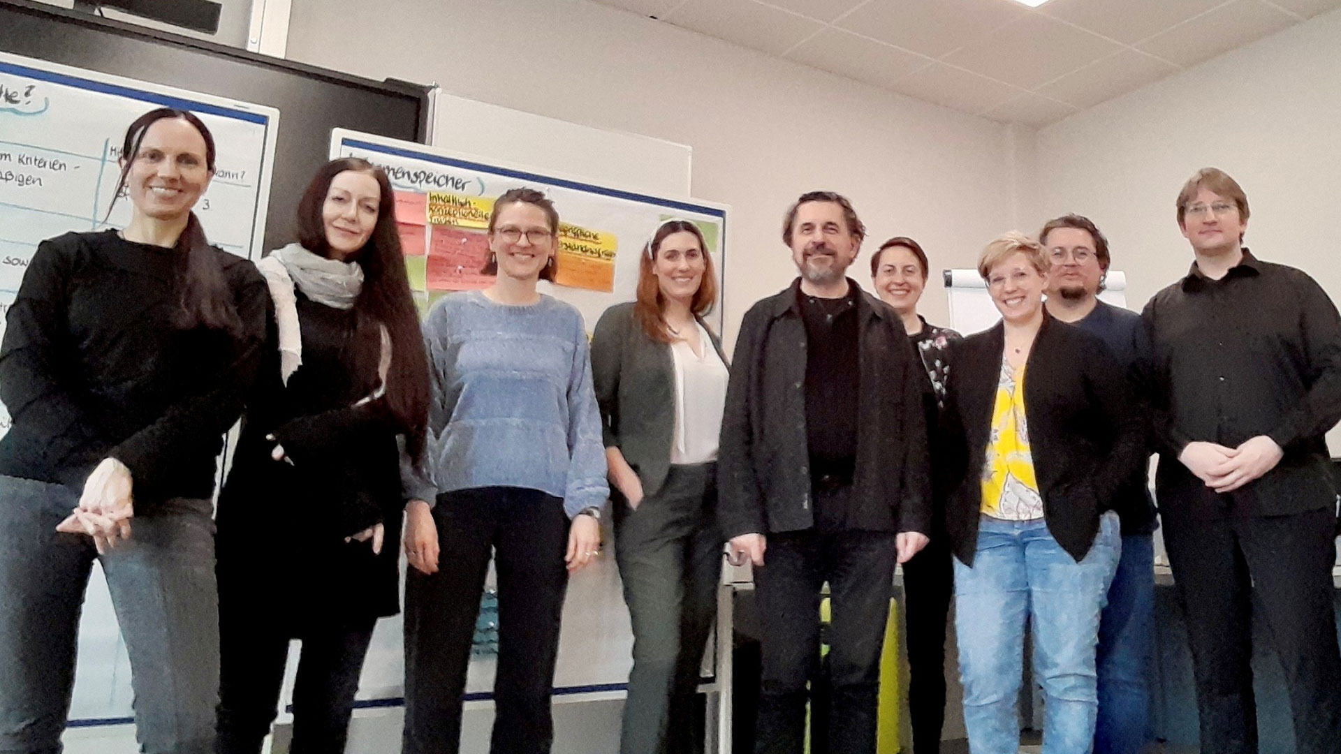 Neun Mitglieder des Projektteams sind beim Kick-Off in Hamburg in einem Seminarraum versammelt und lächeln in die Kamera; im Hintergrund zeigen mit vielen Notizen beklebte Whiteboards ihre bisherige Planungs- und Koordinationsarbeit.
