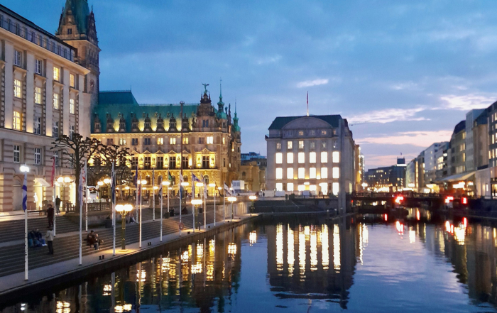 Eine schöne Ansicht der Hamburger Altstadt in der Dämmerung mit Blick aufs beleuchtete Rathaus und der spiegelnden Wasseroberfläche der Kleinen Alster im Vordergrund.
