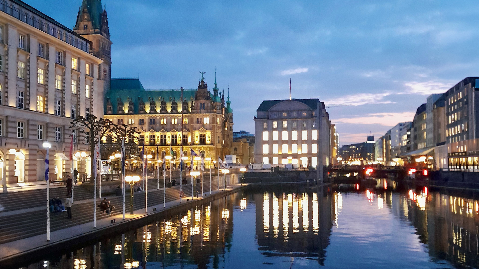 Eine schöne Ansicht der Hamburger Altstadt in der Dämmerung mit Blick aufs beleuchtete Rathaus und der spiegelnden Wasseroberfläche der Kleinen Alster im Vordergrund.
