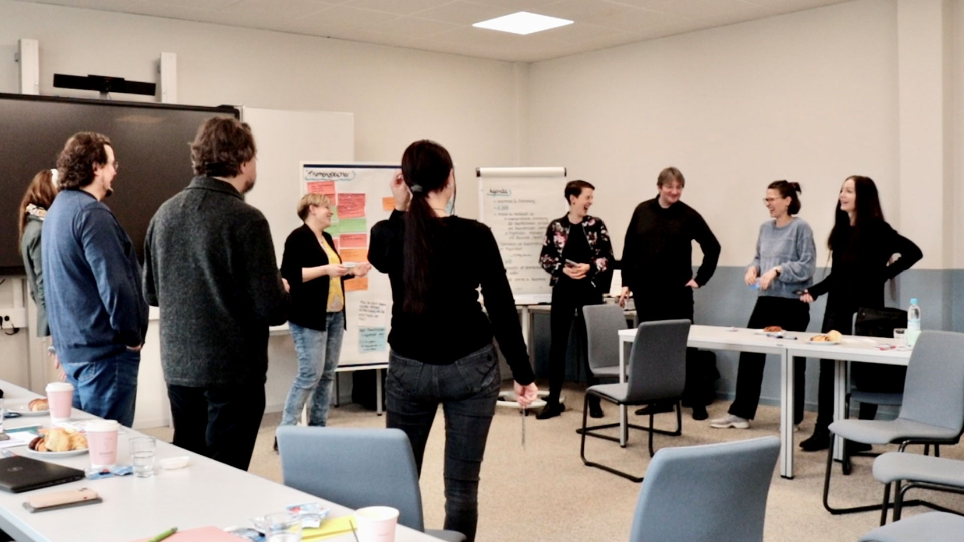 Die neun Projektmitarbeiter, die sich beim Kick-Off in Hamburg getroffen haben, stehen während der Durchführung eines Gemeinschaftsspiels gestikulierend und laut lachend im Seminarraum.